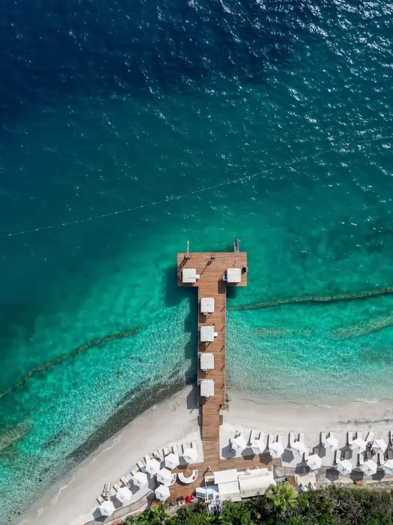Hava fotoğrafında, Göcek'in berrak turkuaz sularına uzanan, üzerinde beyaz masa ve sandalyelerin bulunduğu uzun ahşap bir iskele ile plajda sıra sıra dizilmiş beyaz şemsiye ve şezlonglar, bölgenin sakin ve güzel gezilecek yerlerinden birini gözler önüne seriyor.