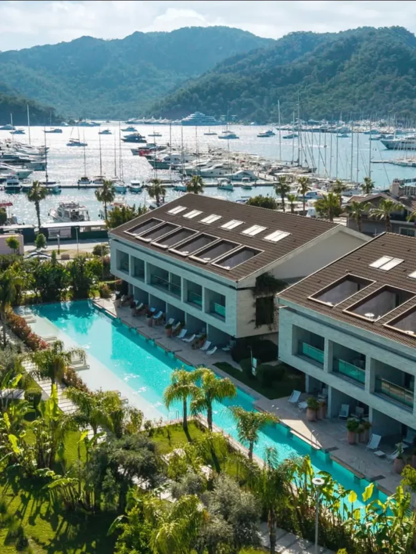 Göcek'in doğal güzellikleri içinde konumlanmış modern bir otel veya tesisi kuşbakışı gösteren bu geniş açılı görüntüde, uzun, berrak mavi bir yüzme havuzu, yemyeşil bahçeler, yatlarla dolu parıldayan bir marina ve görkemli ormanlık dağlar dikkat çekerek buranın gezilecek yerler arasındaki cazibesini ortaya koyuyor.
