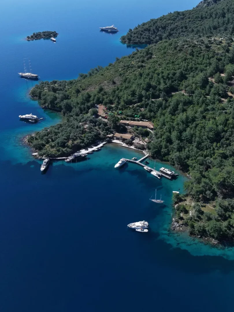 Göcek'in gezilecek yerlerinden eşsiz bir manzara sunan bu havadan çekilmiş fotoğrafta, yemyeşil ormanlarla kaplı kıyılar masmavi ve turkuaz denizin berrak sularıyla buluşurken, birçok lüks yat ve tekne huzur içinde demirlemiş olarak görülüyor.