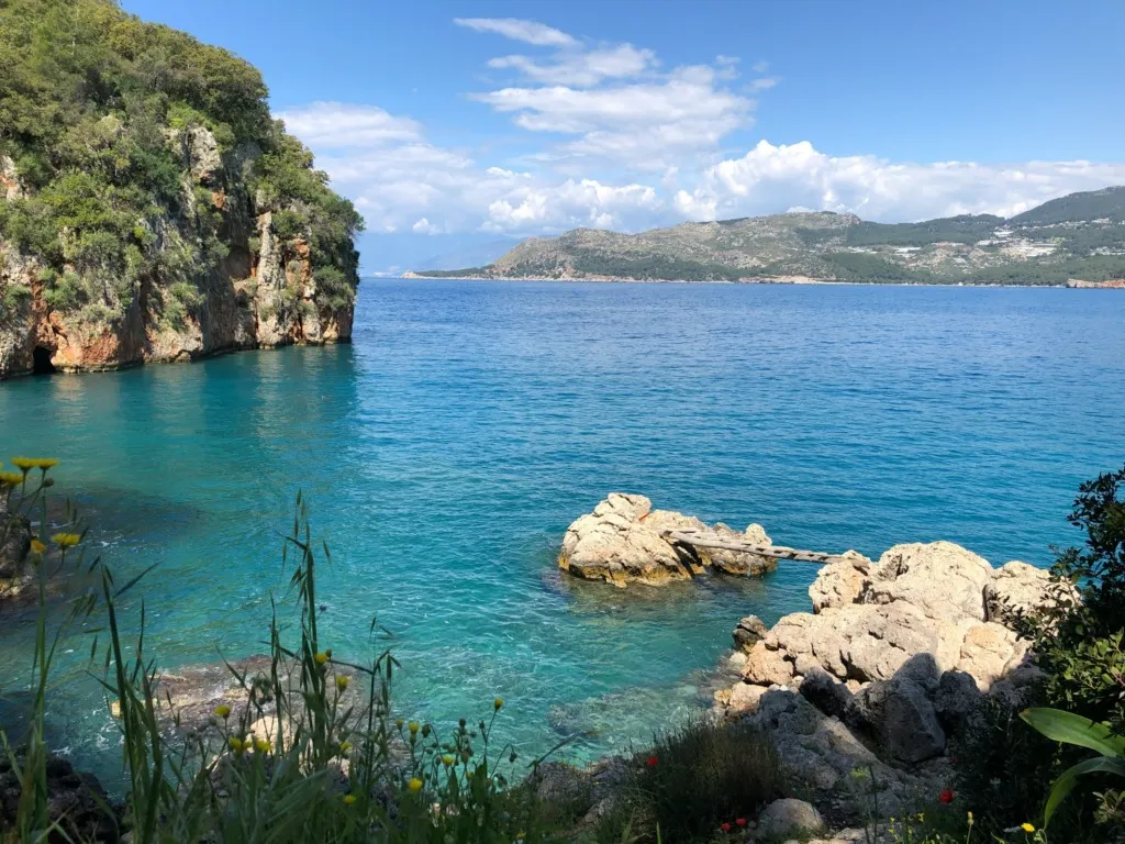 Göcek'in gezilecek yerlerinden biri olan bu büyüleyici koyda, solda yemyeşil ağaçlarla kaplı dik kayalıklar, cam gibi berrak turkuaz renkteki suların içindeki kayalar arasında uzanan küçük bir ahşap köprü ve uzakta yeşil tepeler belirirken, ön planda sarı çiçekler ve çeşitli bitkiler görülüyor.