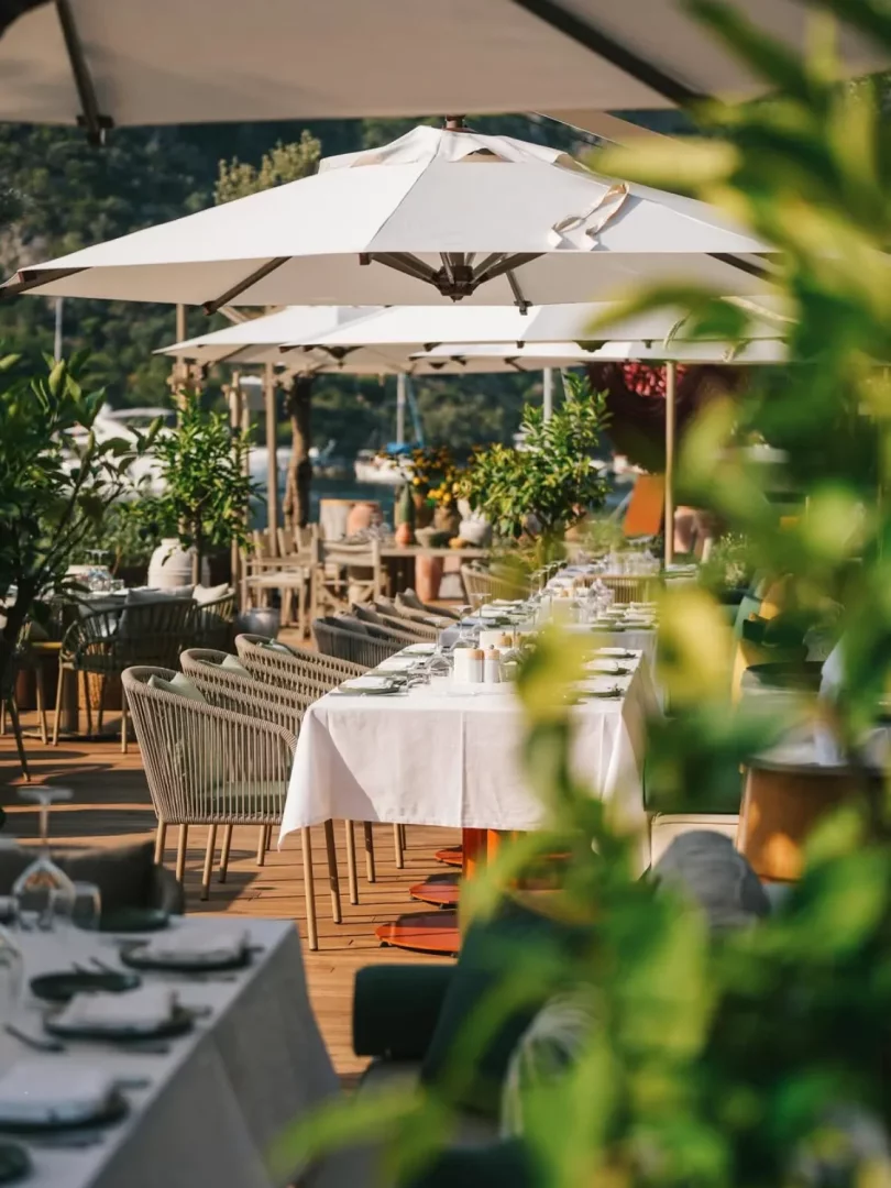 Beyaz şemsiyeler altında, modern hasır sandalyeler ve özenle hazırlanmış masaların sıralandığı, ahşap bir teras üzerindeki açık hava restoran, arka planda marinadaki tekneler ve yemyeşil tepelerle Göcek'in gezilecek yerlerinden birini gözler önüne seriyor.