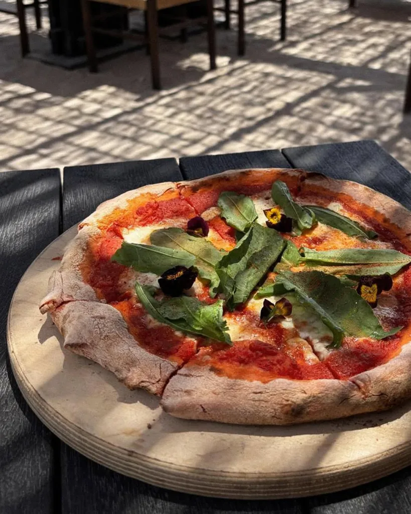 Göcek'te gezilecek yerlerden birinde, koyu renkli ahşap bir masada servis edilen, üzerinde domates sosu, erimiş mozzarella peyniri, taze yeşil roka yaprakları ve mor-sarı yenilebilir çiçeklerle süslenmiş, dış mekanın güneşli ve gölgeli atmosferini yansıtan rustik görünümlü bir pizza, ahşap kesme tahtası üzerinde iştah açıcı bir şekilde duruyor.