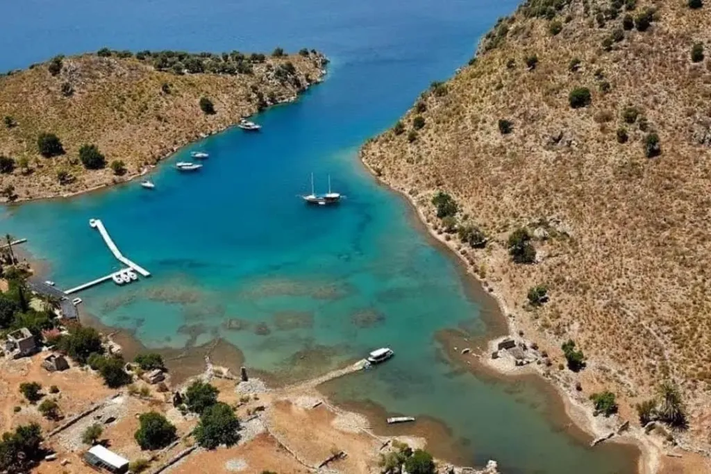 Göcek'in eşsiz doğal güzelliklerini yansıtan bu hava fotoğrafında, turkuazdan derine doğru değişen berrak sularıyla ünlü bir koy görünüyor; kurak tepelerle çevrili koyda yüzen beyaz bir iskele, kıyıya yakın demirli tekneler ve ortada zarif bir yelkenli, buranın popüler bir gezilecek yer olduğunu vurguluyor.