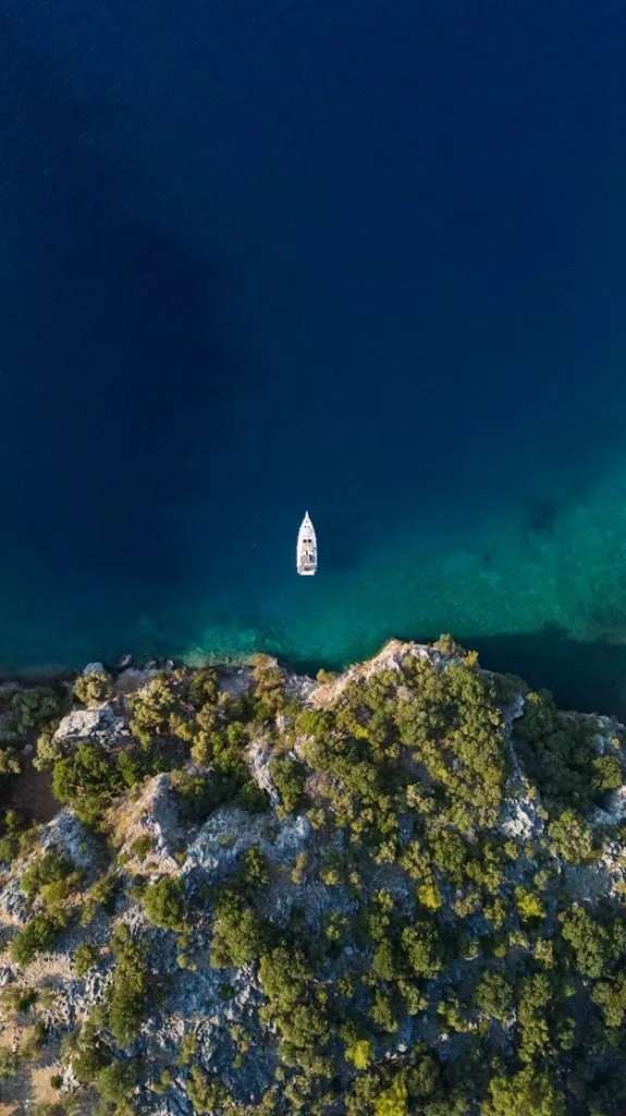 Göcek'in eşsiz doğal güzelliklerini gözler önüne seren bu etkileyici hava fotoğrafında, derin masmavi denizin berrak sularında zarifçe süzülen beyaz bir tekne ve yemyeşil ağaçlarla kaplı kayalık kıyı şeridi huzur veren bir tatil manzarası oluşturuyor.
