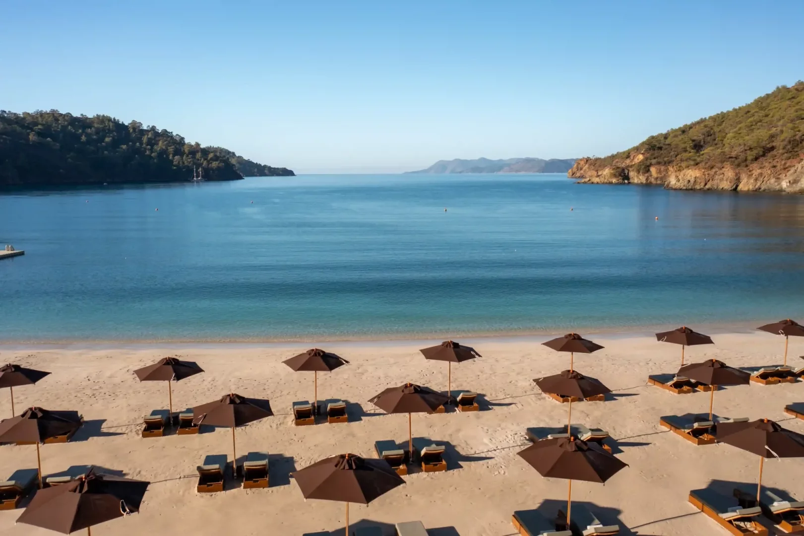 Göcek'in berrak turkuaz sularına sahip sakin koyunda, altın rengi kumlu plaj üzerinde düzenli sıralanmış kahverengi şemsiyeler ve şezlonglar, yemyeşil tepelerle çevrili bu huzurlu gezilecek yerin büyüleyici doğal güzelliğini gözler önüne sermektedir.