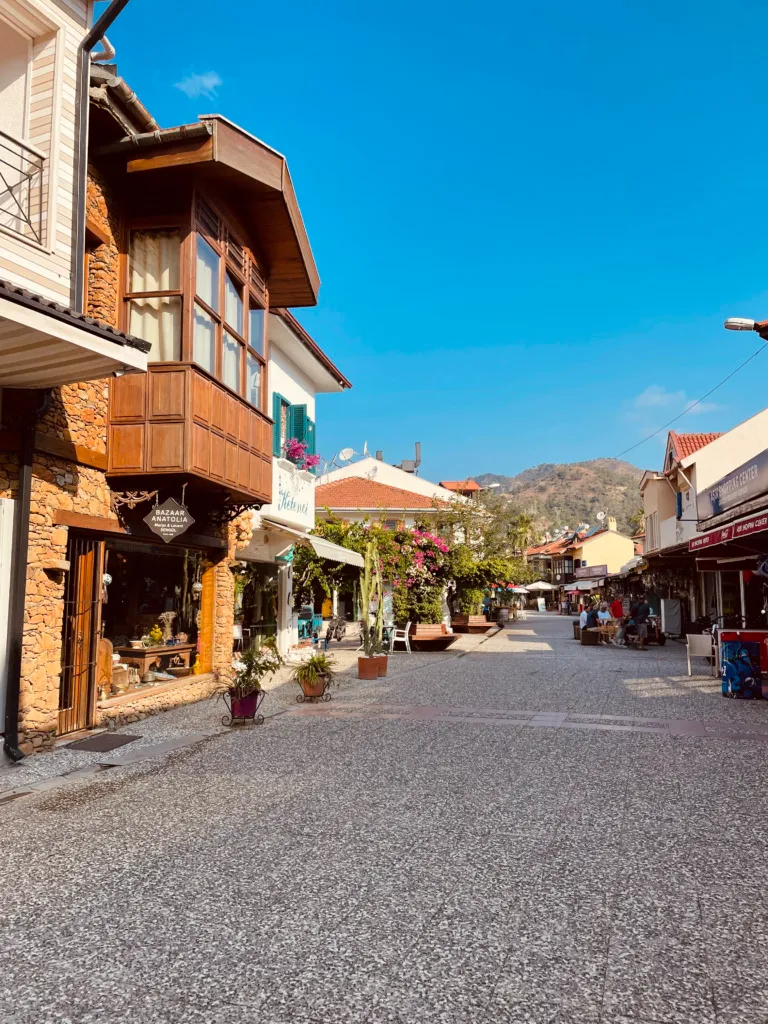 Göcek'in popüler gezilecek yerlerinden biri olan bu Arnavut kaldırımlı cadde; taş cepheli dükkanlar, ahşap cumbalı evler, canlı çiçekler ve uzakta yükselen dağ manzarası eşliğinde, güneşli bir günde keyifli bir yürüyüş imkanı sunuyor.
