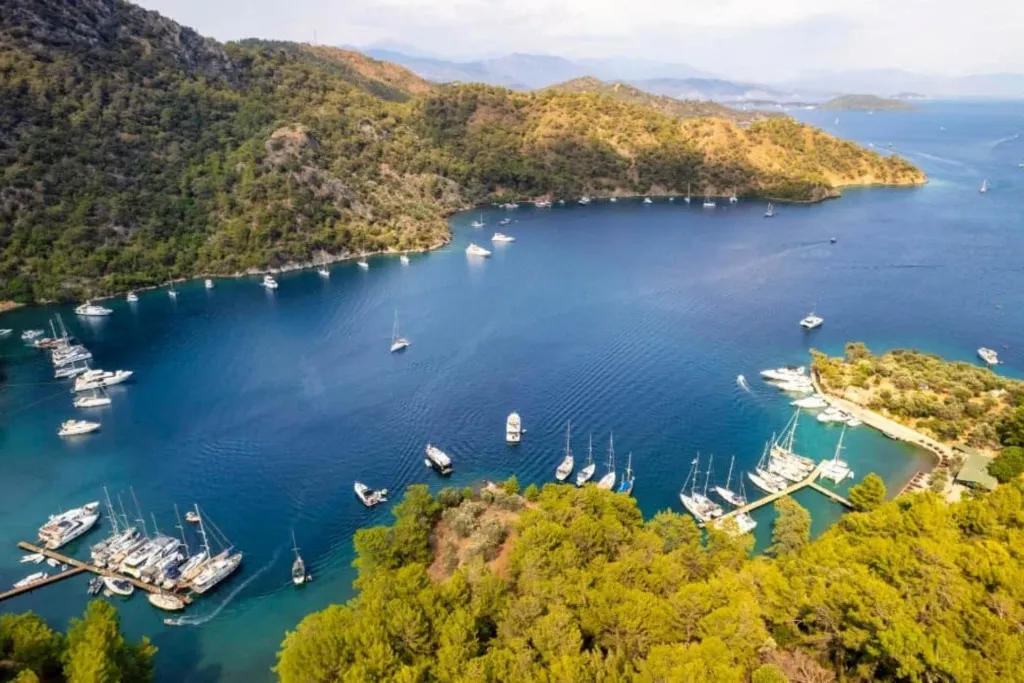 Göcek'teki Bedri Rahmi Koyu'nun havadan çekilmiş muhteşem görüntüsü, zümrüt yeşili çam ağaçlarıyla çevrili masmavi sularda demirlemiş çok sayıda tekne ve yat ile Göcek gezilecek yerler arasındaki doğal güzelliğini sergiliyor.