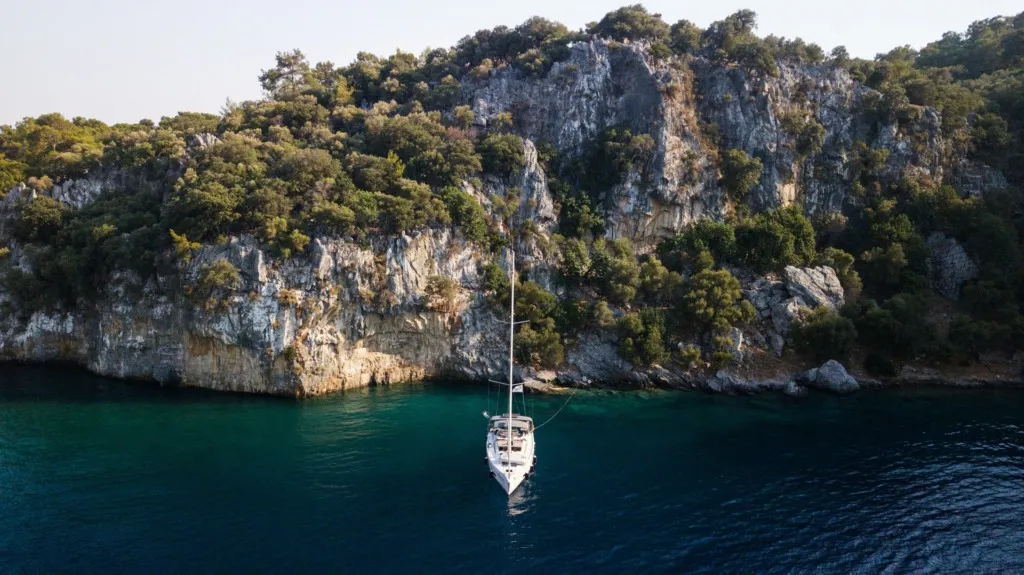 Göcek'in büyüleyici gezilecek yerlerinden biri olan bu huzurlu koyda, yemyeşil ve sık ağaçlarla kaplı yüksek, dik kayalıkların önünde, sakin ve berrak turkuaz sulara demirlemiş zarif beyaz bir yelkenli tekne, tepeden çekilmiş bir açıyla görülüyor.