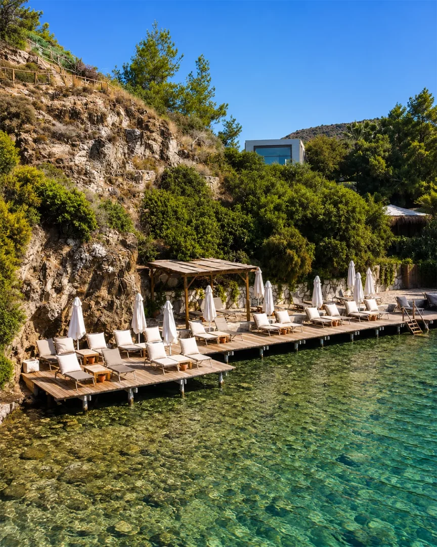 Marmaris Bordubet'in dingin sularına uzanan ahşap iskeleler üzerinde sıralanmış bembeyaz şezlonglar ve güneş şemsiyeleri, berrak turkuaz denizi ve yemyeşil kayalık yamaçlarla çevrili, lüks bir tatil köyü ambiyansı sunan huzurlu bir koy manzarası.