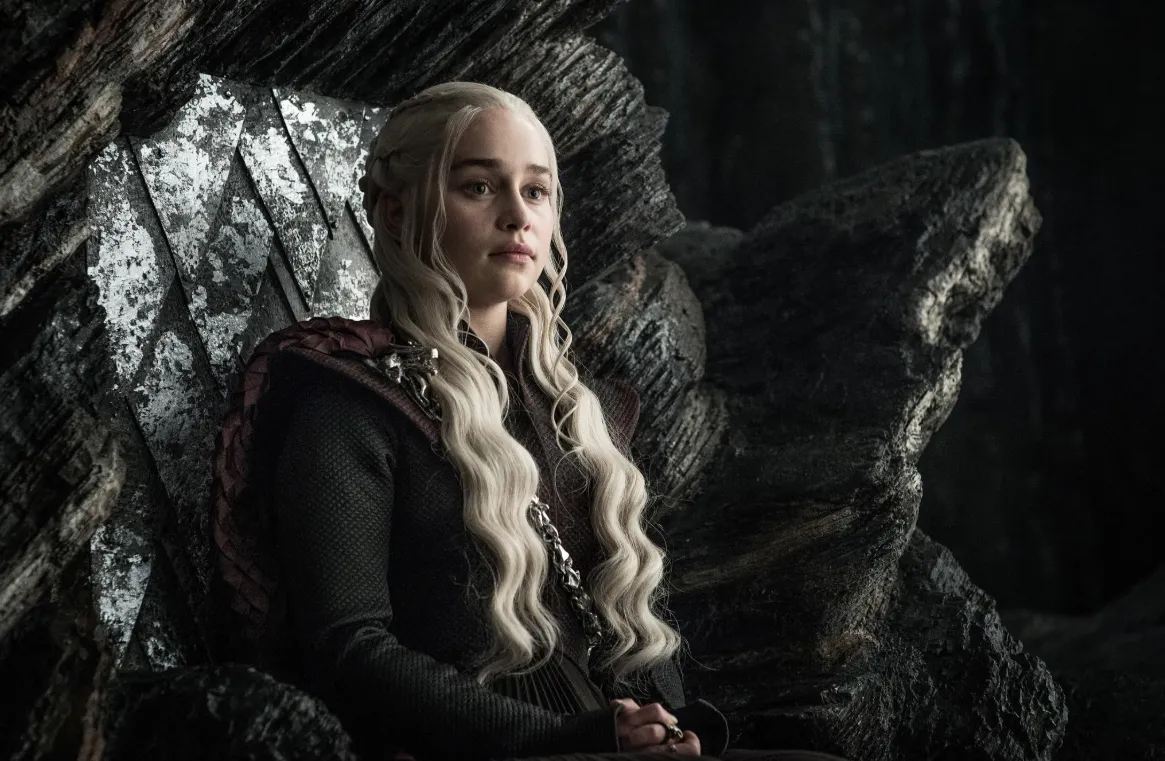 Game of Thrones dizisinden tanıdığımız Daenerys Targaryen, omuzlarında ejderha pulu detayları ve zincirle süslü koyu renkli kıyafetiyle, platin sarısı dalgalı saçları salınmış halde, House of the Dragon'daki atalarına benzer şekilde koyu taşlardan oyulmuş taht benzeri bir koltukta, düşünceli ve kararlı bir ifadeyle oturuyor.