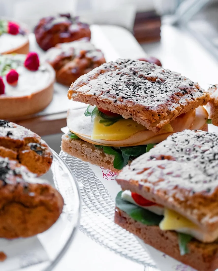 Ahşap masa ve sandalyelerin, duvarları süsleyen yeşil bitkilerin ve süzülen doğal ışığın yarattığı samimi ve davetkar bir atmosfere sahip Grandma Cafe'nin kalabalık iç mekanında, sohbet eden ve kahvelerini yudumlayan çeşitli müşteriler rahat bir ortamda görülüyor.