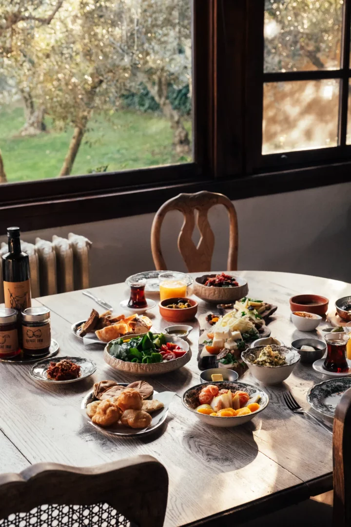 Şirince'deki Güllü Konakları'nda, pencereden görünen zeytin ağaçları eşliğinde, taze yeşillikler, peynir çeşitleri, zeytin, pişi, reçeller ve sıcak çaylar ile donatılmış rustik ahşap masa üzerinde sunulan zengin bir köy kahvaltısı.