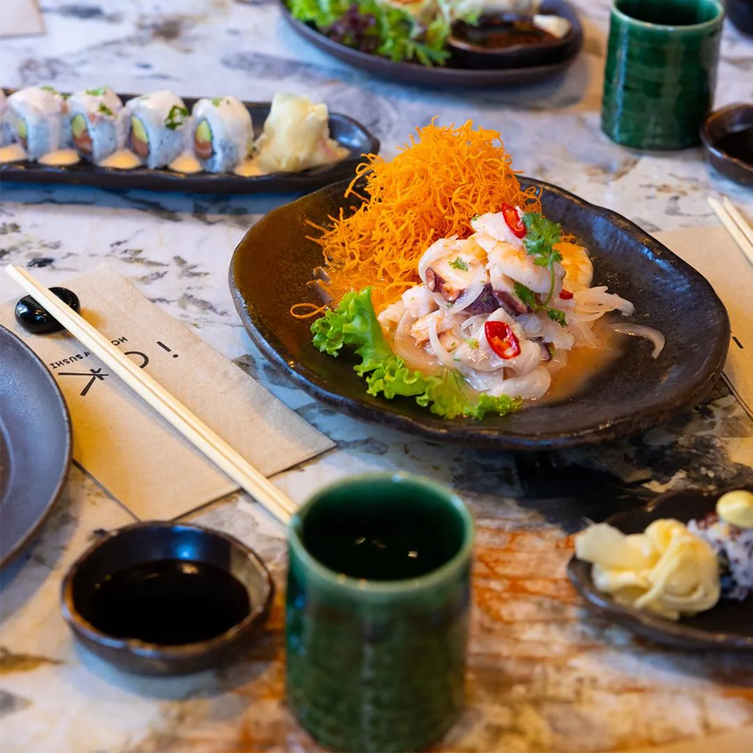 Mermer desenli bir masanın üzerinde, taze deniz mahsulleri salatası ve çıtır turuncu ipliklerle zenginleştirilmiş koyu renkli bir ana yemek tabağı, arka planda kremalı soslu sushi ruloları ve zencefil turşusu içeren başka bir sunum tabağı, ön planda ise bir çift yemek çubuğu, küçük bir soya sosu kasesi ve yeşil seramik içecek bardakları düzenli bir şekilde konumlanmıştır.