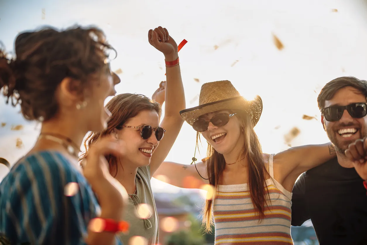 Güneşli havada neşeli bir festival anında, yüzlerinde kahkahalarla dolu ifadeler ve gözlerinde şık güneş gözlükleriyle, konfeti yağmuru altında kolları havada dans edip coşkulu bir kutlama yapan dört genç arkadaş.