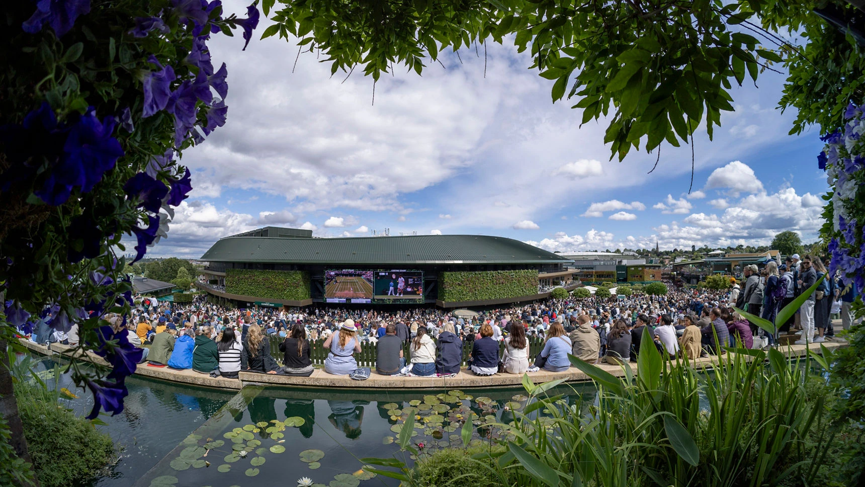 Wimbledon Tenis Turnuvası sırasında, Henman Hill'deki (şimdiki adıyla Murray Mound) dev ekranda bir tenis maçını izleyen coşkulu kalabalık, ön planda nilüferli bir gölet ve mor çiçeklerle çerçevelenmiş canlı İngiliz yaz atmosferini gözler önüne seriyor.