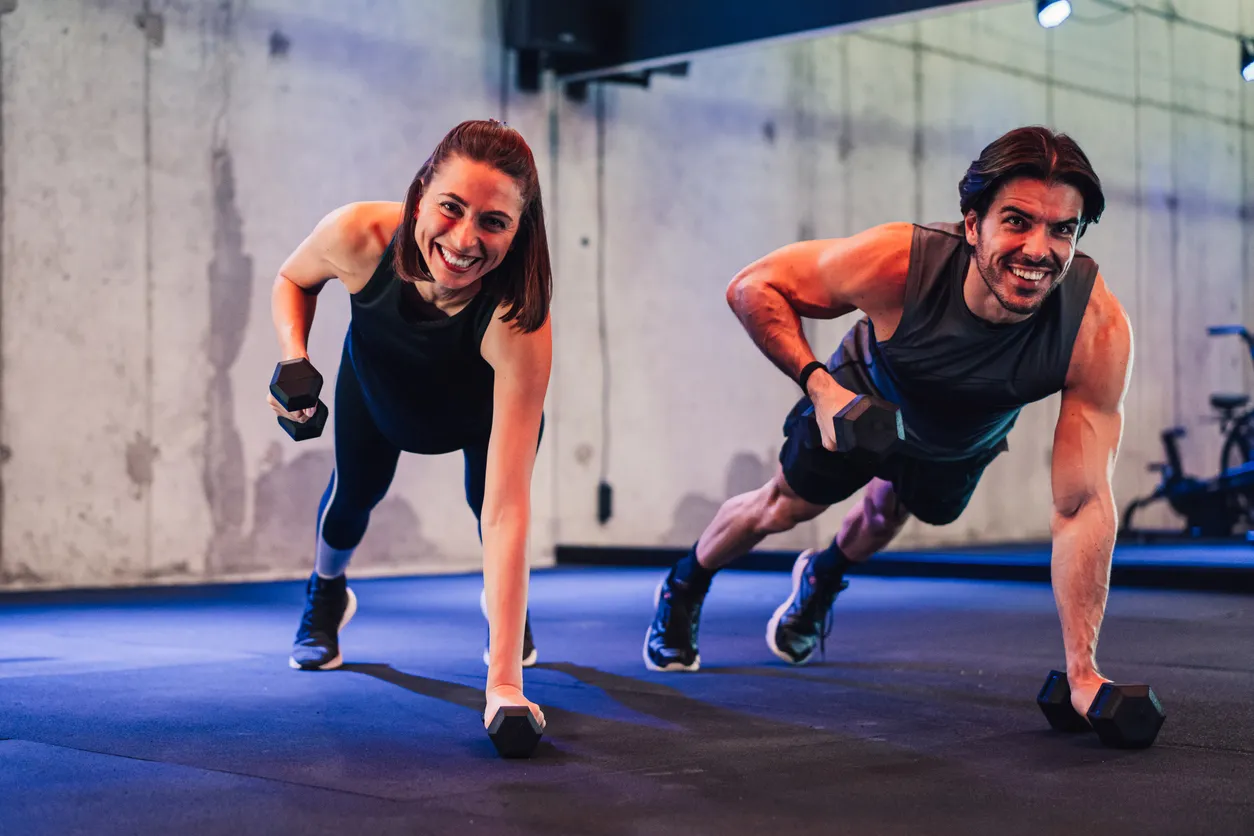 Modern bir spor salonunda, plank pozisyonunda dambıl ile kürek çekme hareketi yapan ve kameraya neşeyle gülümseyen bir kadın ile bir erkek, HIIT kardiyo antrenmanının dinamik ve zorlayıcı doğasını canlı bir şekilde sergiliyor.