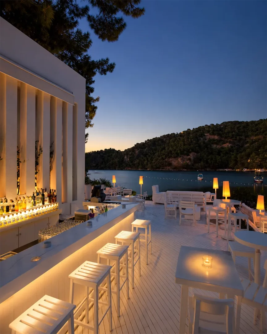 Fethiye'deki Hillside Beach Club'ın bembeyaz ahşap zeminli terasında, denize ve yemyeşil tepelere nazır modern bir açık hava barı, akşam alacakaranlığında sıcak ışıklarla aydınlatılmış tezgahı ve beyaz oturma gruplarıyla sakin bir atmosfer sunuyor.