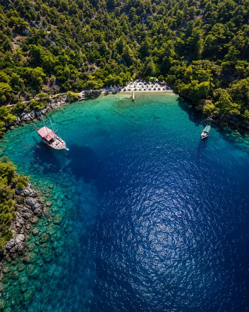 Fethiye'deki yemyeşil tepelerle çevrili, berrak turkuaz sulara sahip özel bir koyun havadan görünümü, şezlong ve şemsiyelerle dolu plajı, iskelesi ve demirli tekneleriyle Hillside Beach Club'ın huzurlu atmosferini yansıtmaktadır.