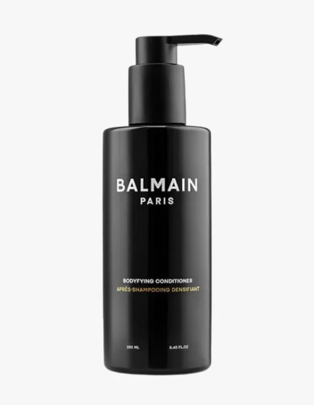 Düz beyaz bir zeminde sergilenen, Balmain Paris markasına ait, üzerinde BODYFYING CONDITIONER yazan siyah renkli, pompalı bir şişe, özellikle erkek saçlarına hacim ve dolgunluk katmak üzere tasarlanmış bu dolgunlaştırıcı saç kremini net bir şekilde gösteriyor.
