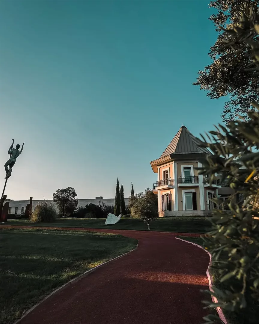 Eceabat'taki Hotel Caeli'nin geniş yeşil bahçesinde, sağda kendine özgü kule çatılı ve balkonlu açık sarı renkli ana bina, önünde kavisli kırmızı yürüyüş yolu, sol tarafta dik bir heykel, uzakta beyaz soyut bir kuş heykeli ve sağ üstten kadraja giren zeytin ağacı dallarıyla masmavi gökyüzü altında huzurlu bir manzara sergileniyor.