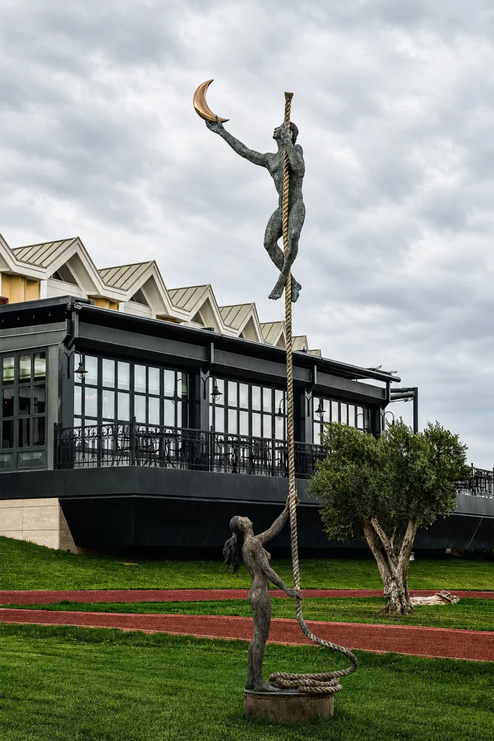Bulutlu bir gökyüzü altında, Hotel Caeli'nin kendine özgü çatılı modern binasının önündeki yeşil çimenlik alanda, ip üzerinde yükselip altın bir hilali yakalamaya çalışan erkek figürü ve ipin dibinde yukarı bakan bir kadın figürünün yer aldığı bronz heykel.