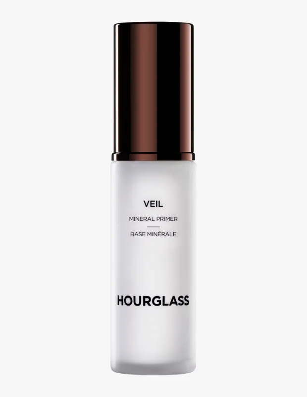 Mat beyaz, buzlu görünümlü silindirik şişesi ve parlak koyu kahverengi kapağıyla dikkat çeken, üzerinde büyük harflerle HOURGLASS ve VEIL MINERAL PRIMER yazıları okunan bir makyaj bazı ürünü fotoğrafı.