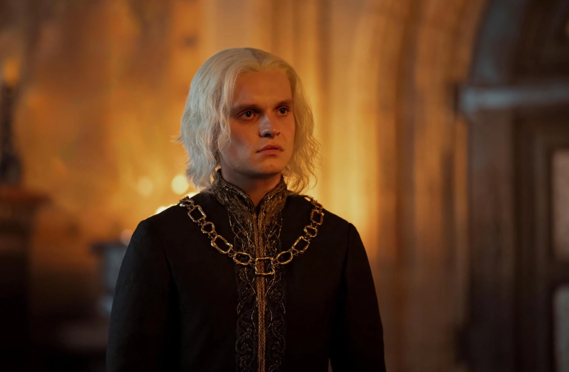 House of the Dragon dizisinin önemli karakterlerinden Kral II. Aegon Targaryen, iç savaşın getirdiği taht mücadelesinin ağırlığını taşırcasına, gümüşi platin renkli uzun saçları, yüzündeki düşünceli ifadesi ve altın detaylı siyah, görkemli kıyafetiyle kadraja yansıyor.
