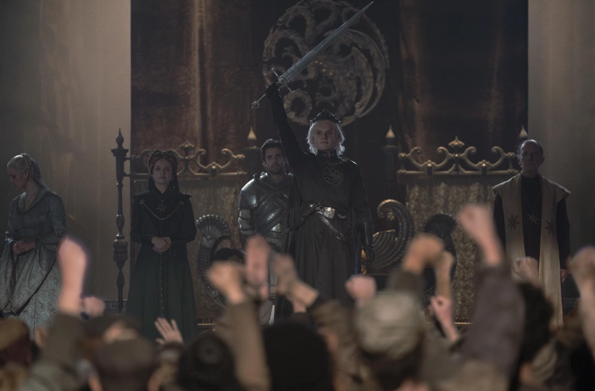 House of the Dragon dizisinden bir sahnede, Kral II. Aegon Targaryen taç giyme töreninde kılıcını havaya kaldırırken, arkasında annesi Alicent Hightower ve ablası Helaena ile birlikte coşkulu kalabalığı selamlıyor, bu an Demir Taht için süregelen kanlı taht mücadelesinin başlangıcını simgeliyor.