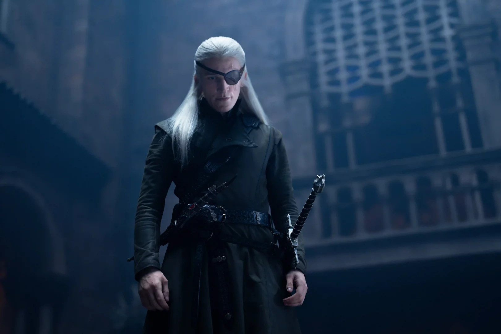 Aemond Targaryen, House of the Dragon'ın 3. sezonunda, beyaz saçları, sol gözündeki karanlık göz bandı ve belinde kılıcıyla Ejderhaların Dansı'ndaki amansız mücadelesini yansıtan kararlı bir duruşla, loş ışıklı bir salonda kameraya bakıyor.