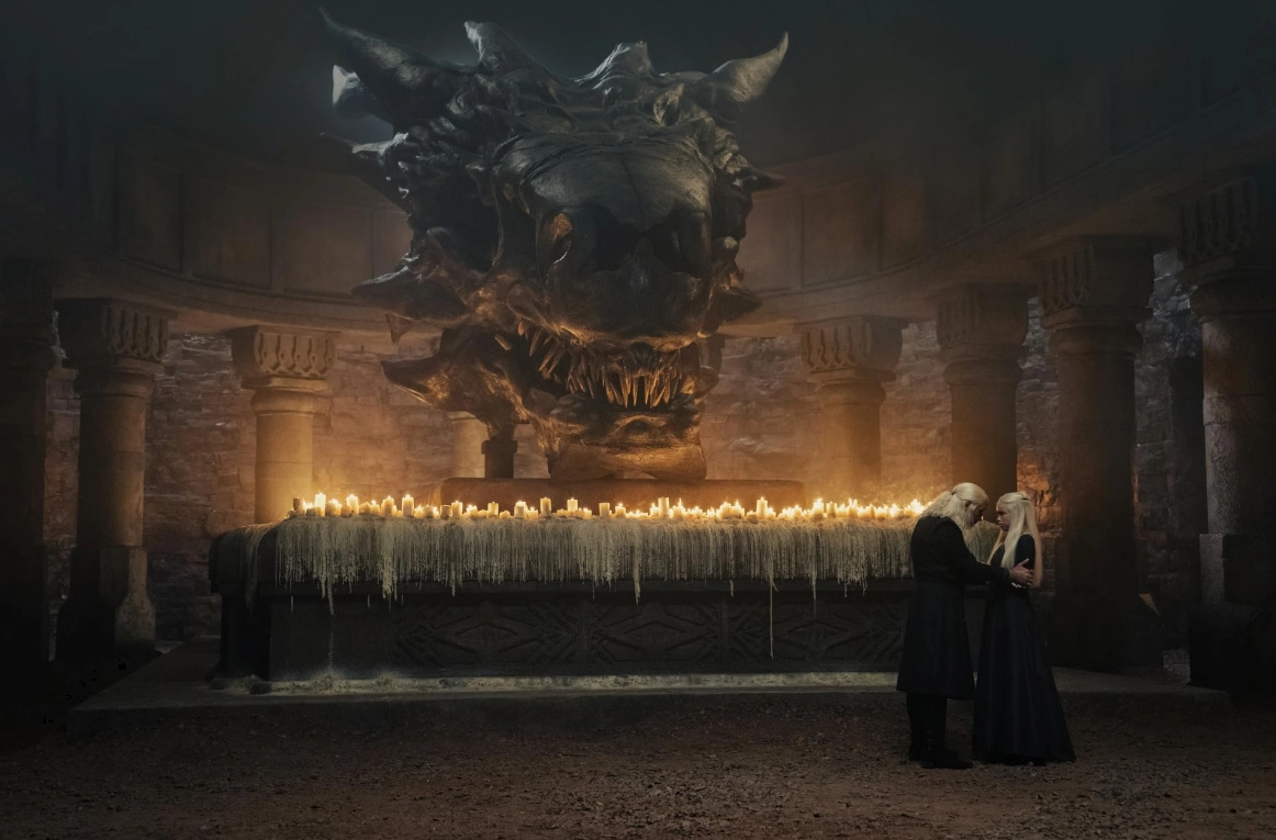 House of the Dragon dizisinden bir sahnede, beyaz saçlı bir erkek figürü, mumlarla bezeli uzun bir sunağın arkasında duran Balerion'ın devasa ejderha kafatasının altında, loş ışıklı bir mahzende, beyaz saçlı bir kadına sarılarak onu teselli ediyor.