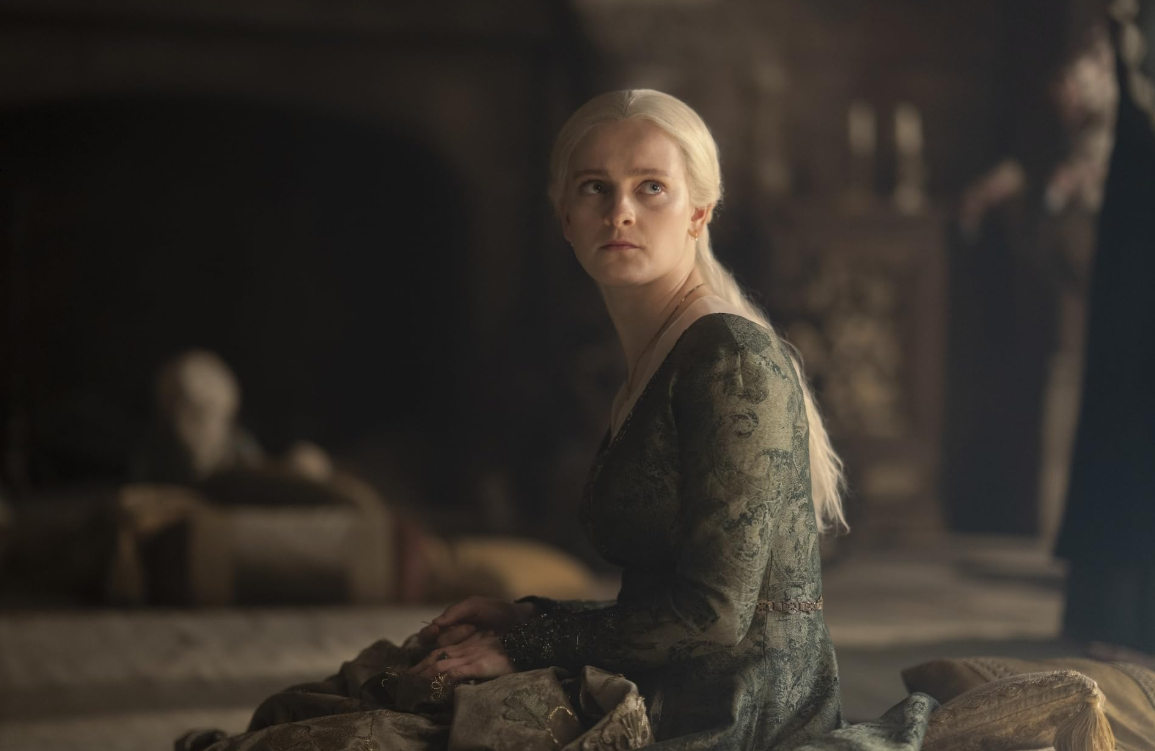 House of the Dragon 3. Sezon'dan bir sahnede, uzun platin sarı saçlı Helaena Targaryen, desenli koyu yeşil elbisesiyle otururken, aile bağları ve siyasi ittifakların karmaşıklaştığı bu dönemde, derin bir düşünceyle hafifçe yukarı bakarak adeta Ejderhaların Dansı'nın geleceğine dair şifreli kehanetlerini dillendirecek gibi duruyor.
