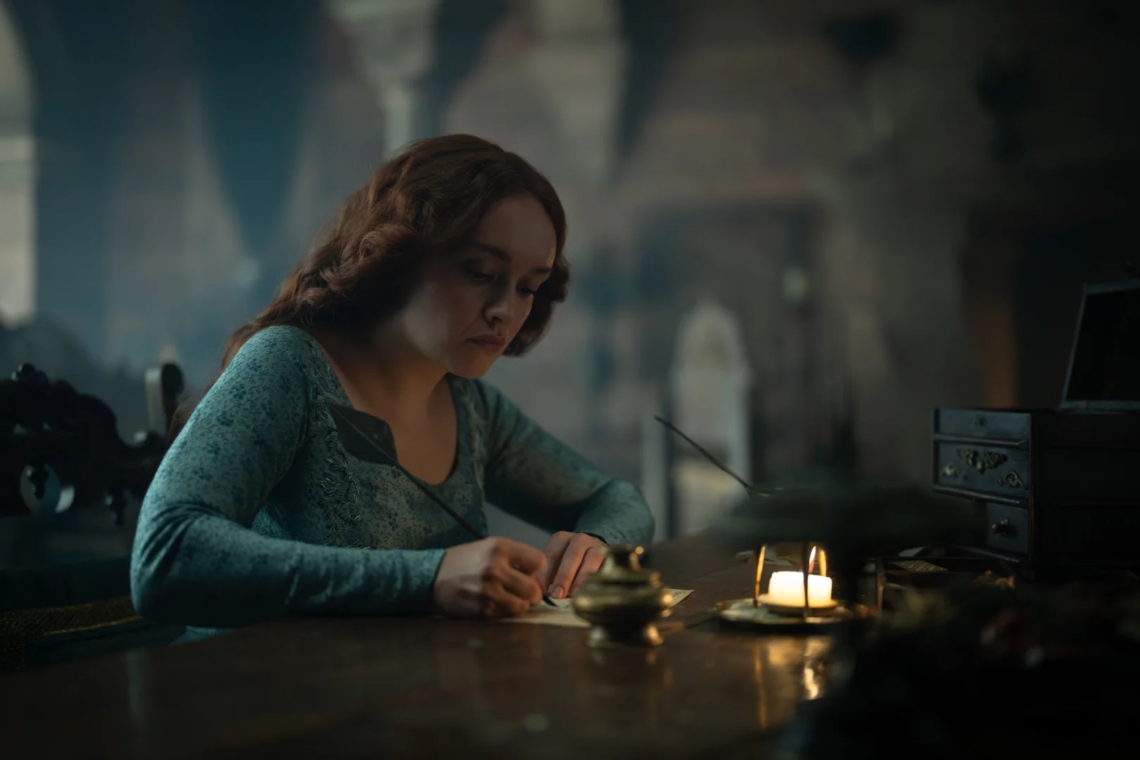House of the Dragon dizisinden Alicent Hightower rolündeki Olivia Cooke, loş ışıklı bir odada ahşap masasında mum ışığında tüy kalemle dikkatle bir şeyler yazarak, Westeros'un taht mücadelesinde Yeşiller'in siyasi liderlerinden biri olarak stratejilerini belirliyor.
