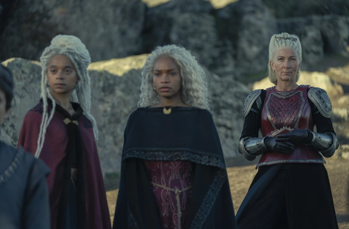 House of the Dragon dizisinden üç karakter, gri kayalık bir zeminde, gümüş saçlarıyla dikkat çekiyor: zırhlı ve vakur Prenses Rhaenys Targaryen, orta planda siyah pelerinli ve altın kolye uçlu Baela Targaryen ile solda bordo pelerinli ve daha genç görünen ikizi Rhaena Targaryen, Ejderhaların Dansı'ndaki rollerini bekler gibi ciddi bir ifadeyle duruyor.