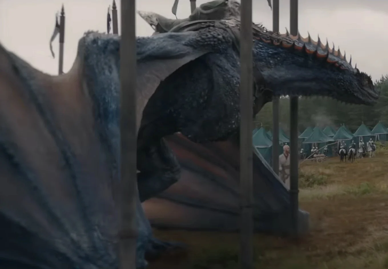 House of the Dragon 3. sezonunda Yeşillerin stratejik gücü olarak ön plana çıkan, parlak bakır renkli kanatlara ve koyu mavi pullara sahip devasa ejderha Tessarion, arkasında yeşil çadırlardan oluşan bir ordu kampı ve atlı askerler görünürken, boynundaki turuncu dikenli sırt çıkıntılarıyla heybetli bir şekilde duruyor, yanında Prens Daeron Targaryen olduğu düşünülen açık renkli giysili bir figür yer alıyor.