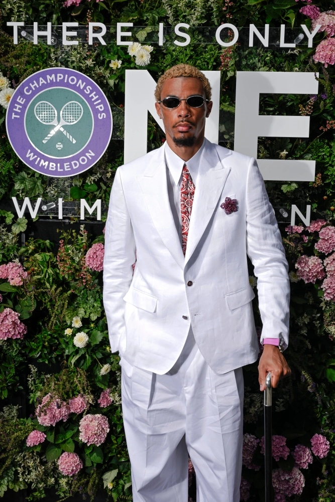 Wimbledon etkinliğinde çiçekli yeşil duvar önünde beyaz takımı, kırmızı desenli kravatı, güneş gözlükleri ve dikkat çeken pembe bantlı Chopard Alpine Eagle 41 Beach Edition kol saatiyle poz veren Hugo Ekitike.