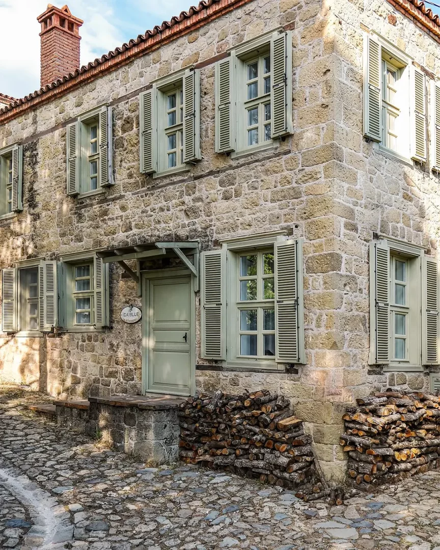 Adatepe köyünde yer alan İda Blue adlı taş evin dış cephesi, ahşap yeşil panjurlu pencereleri, kırmızı kiremit çatısı ve duvara yaslanmış düzenli odun yığınlarıyla pitoresk bir görünüm sergilerken, arnavut kaldırımı sokak dokusuyla bütünleşiyor.