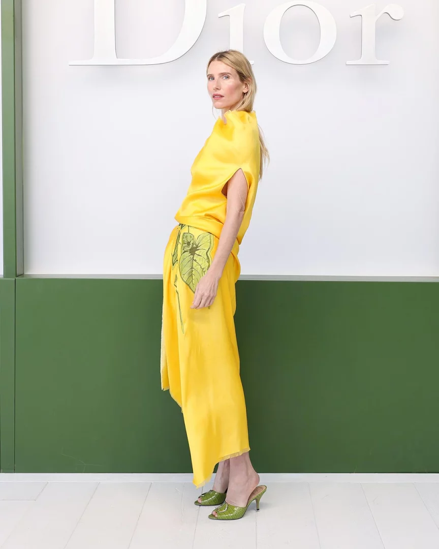Dior logosu ve koyu yeşil bir panelin önünde yan duran, uzun sarı saçları ve sarı renkteki zarif, bol kesim bluzu ile yaprak desenli etek giyen bir kadın, tamamlayıcı yeşil topuklu terlikleriyle ilham veren sarı saç görünümünü sergiliyor.