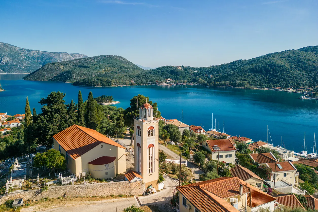 Parlak güneşli bir günde İthaki Adası'nın masmavi körfezini kuşatan yemyeşil tepeler arasında, kırmızı kiremit çatılı evlerin, belirgin bir çan kulesi ile kilisenin ve kıyıda demirli beyaz yelkenlilerin görüldüğü pitoresk bir sahil kasabasının kuşbakışı görüntüsü.