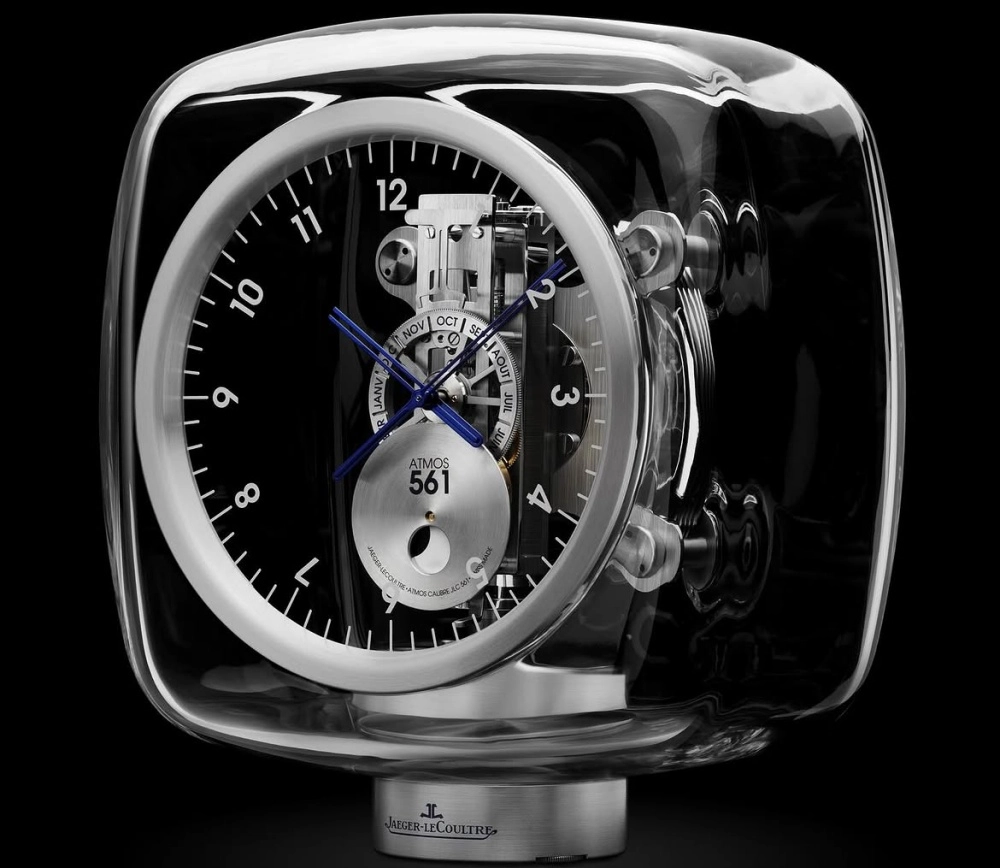 Şeffaf cam gövdesi içindeki görünen karmaşık mekanizması, belirgin mavi akrep ve yelkovanı, ay fazı ile ayları gösteren takvim kadranına sahip, Marc Newson tasarımı modern Jaeger-LeCoultre Atmos 561 masa saati.