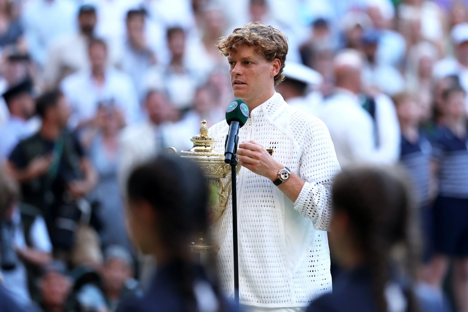 Kıvırcık kızıl saçlı, beyaz fileli bir ceket giyen Jannik Sinner, Wimbledon logolu mikrofon başında, kupasıyla konuşurken, sarı altın Rolex Cosmograph Daytona Oysterflex kayışlı lüks kol saati bileğinde belirgin bir şekilde parlıyor.