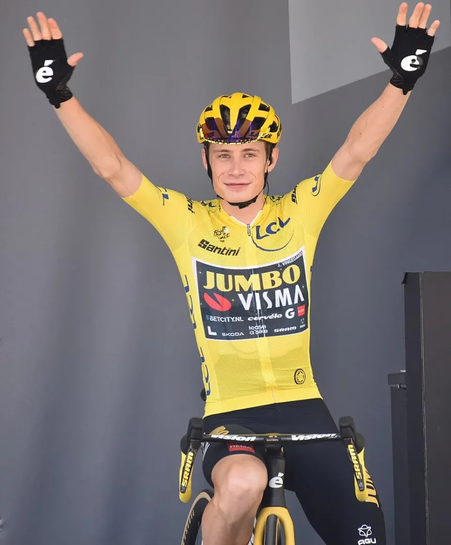 Tour de France 2023 kapsamında sarı mayosuyla bisikleti üzerinde kolları havada zaferini kutlayan Jonas Vingegaard, parlak sarı kaskı ve siyah eldivenleriyle kameraya doğru hafifçe gülümsüyor.