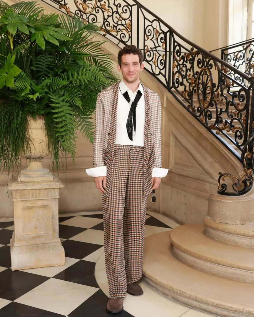 Aktör Josh O'Connor, Dior Sonbahar-Kış 2026 Couture defilesine katılırken, ekose takım elbisesi, beyaz gömleği ve siyah kravatıyla şık bir şekilde duruyor, arkasında görkemli merdiven ve yemyeşil bitkilerle çevrili Paris Haute Couture Moda Haftası'nın lüks ambiyansını yansıtıyor.