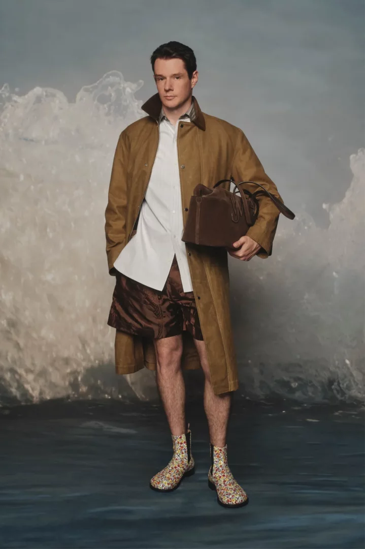 JW Anderson İlkbahar 2027 koleksiyonundan bir model, dalgalı su temalı bir arka planın önünde ayakta durarak, süet benzeri deve tüyü rengi uzun bir kaban, beyaz gömlek, parlak kahverengi şort, elinde koyu kahverengi bir çanta ve rengarenk çiçek desenli botlar giyiyor.