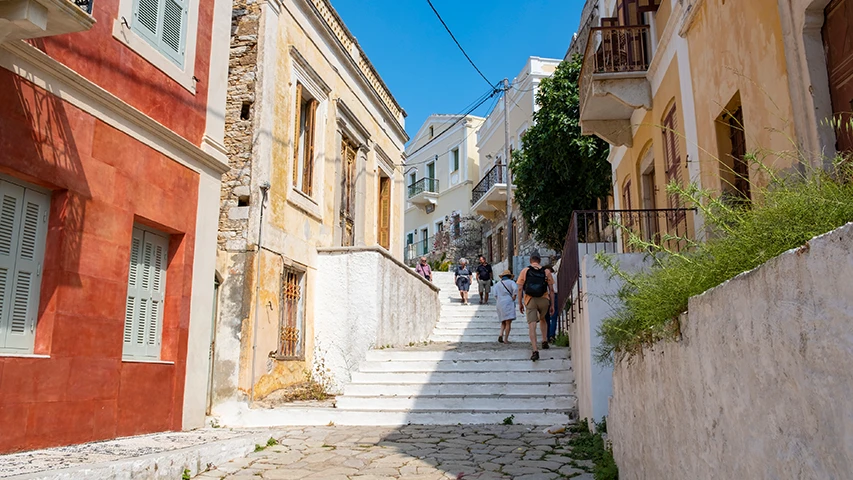 Kali Strata merdivenleri, güneşli bir günde, daracık sokakta yükselen kırmızımsı ve bej renkli eski Rum evlerinin arasından yukarı doğru tırmanan birkaç turistin görüntülendiği, taş döşeli zeminli, otantik bir ada yerleşimi atmosferi sunuyor.