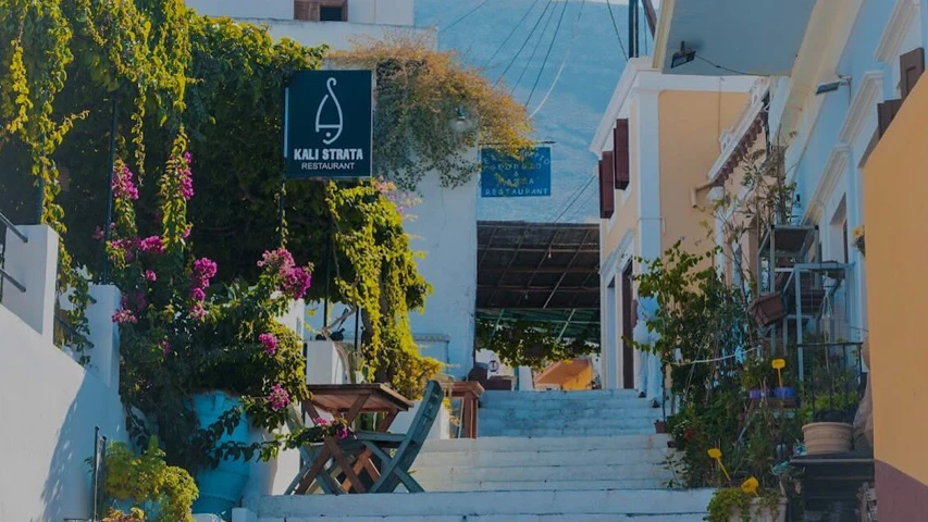 Yemyeşil bitkilerle, mor begonvillerle ve Kali Strata Restaurant tabelasıyla çevrili beyaz ve açık sarı binaların arasından yukarı kıvrılan taş merdivenler, Simi adasının otantik güzelliğini ve canlı atmosferini yansıtıyor.