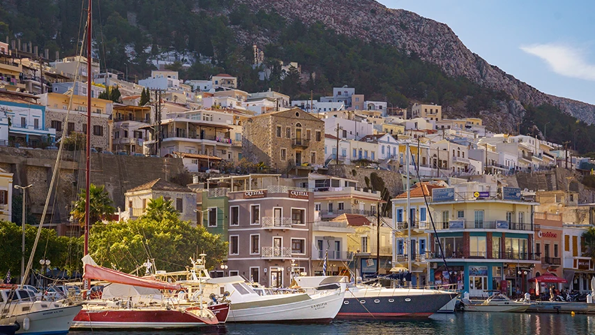 Kalymnos'un hareketli limanında, sarp yamaçlara kurulu, beyaz ve pastel tonlardaki çok katlı binaların arasında, farklı boyutlardaki yelkenli ve motorlu teknelerin berrak sularda demirlediği, adaya nasıl gidileceğini düşündüren pitoresk bir ada yerleşimi manzarası.