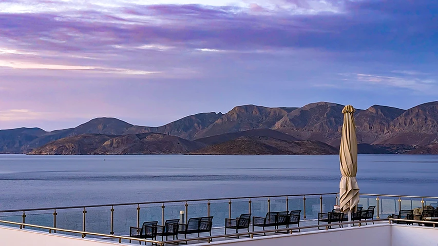 Şafak vakti veya alacakaranlıkta mor ve pembe tonlarındaki gökyüzünün altında, Kalimnos'ta konaklama için ideal olabilecek, denize ve karşıdaki dağlara bakan bir otel balkonundan geniş bir manzara; terasta katlanmış bir şemsiye ve boş oturma grupları görülüyor.