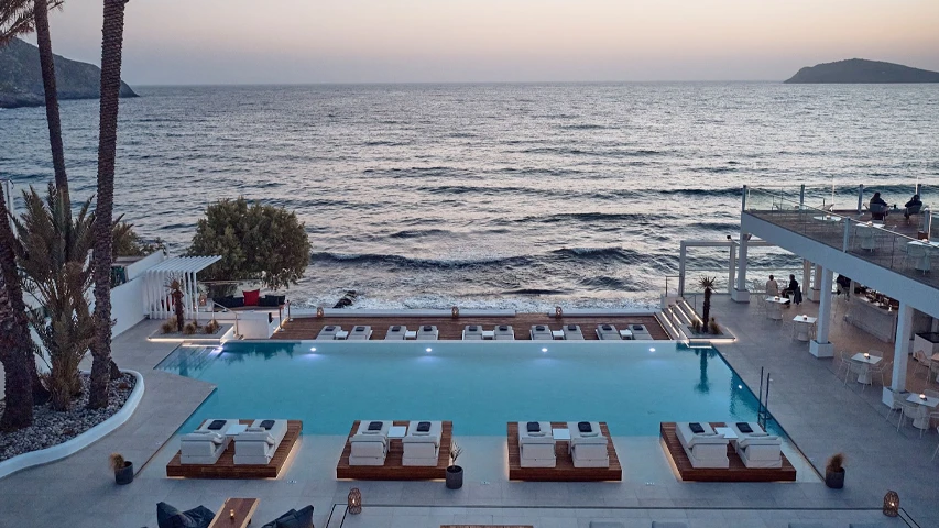 Kantouni Beach Boutique Hotel'in Kalymnos'taki denize nazır modern beyaz mimarisi, dalgalı Ege Denizi manzaralı sonsuzluk havuzunda yüzen konforlu şezlonglar ve teraslardaki şık oturma grupları ile sakin bir akşamüstünü betimleyen geniş bir görüntü.