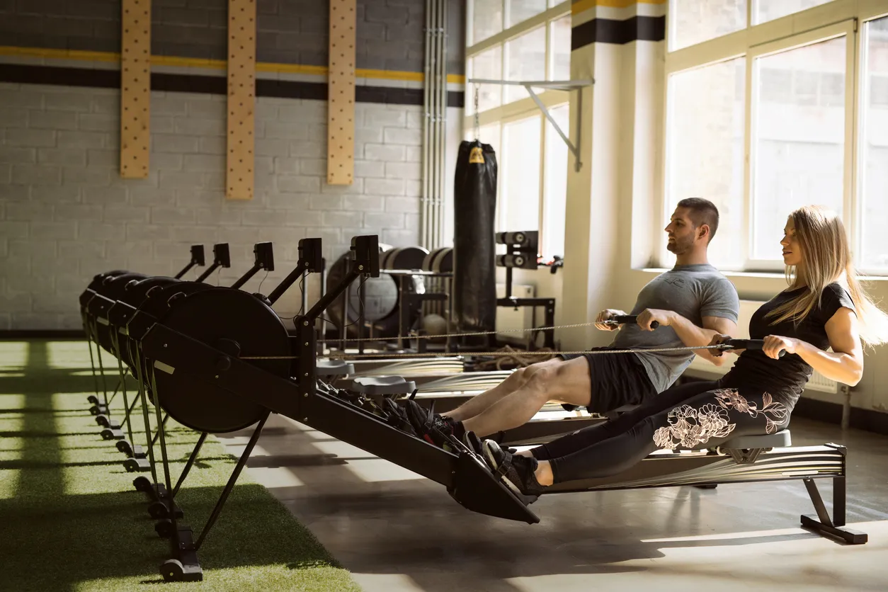 Spor salonunda sıralı kürek çekme makinelerinde senkronize bir şekilde kardiyo egzersizi yapan bir kadın ve bir erkek, kaslarını çalıştırarak sağlıklı yaşamın faydalarını deneyimliyor, enerjileri yüzlerine yansıyor.