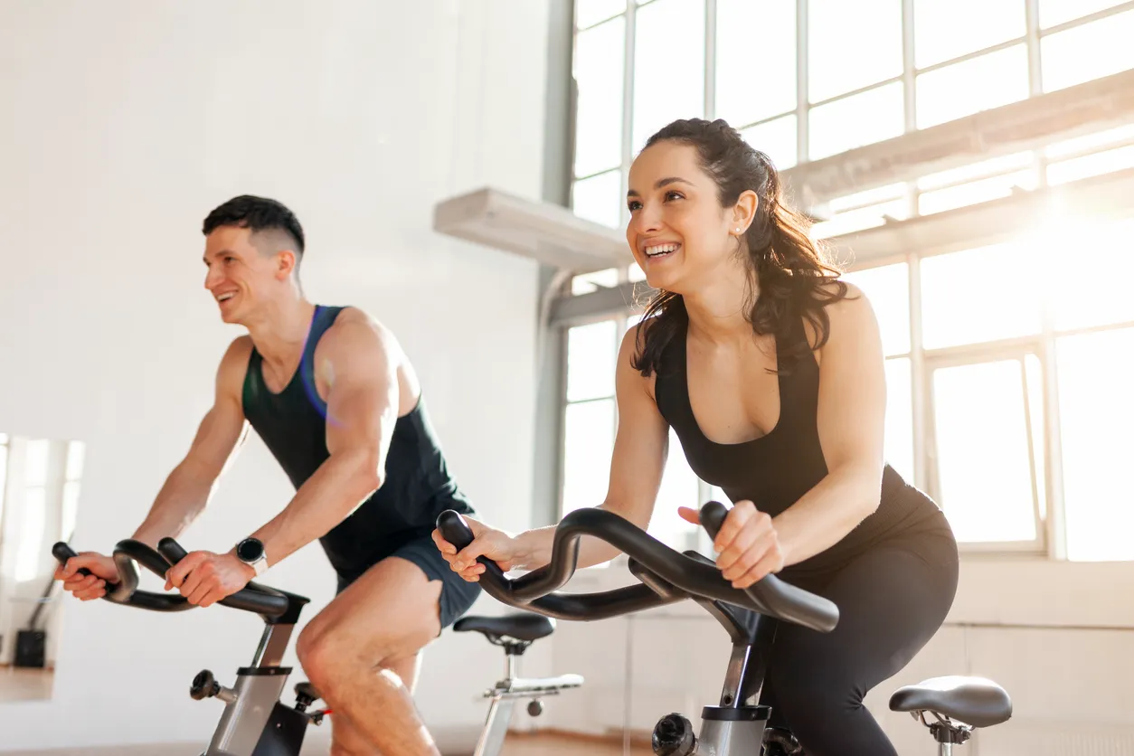 Parlak ışıklı bir spor salonunda, enerjik ve gülümseyen bir kadın ve bir erkek, modern kondisyon bisikletleri üzerinde keyifli bir şekilde kardiyo antrenmanı yaparken görülüyor, arka plandaki geniş pencerelerden gelen doğal ışık mekanı aydınlatıyor.